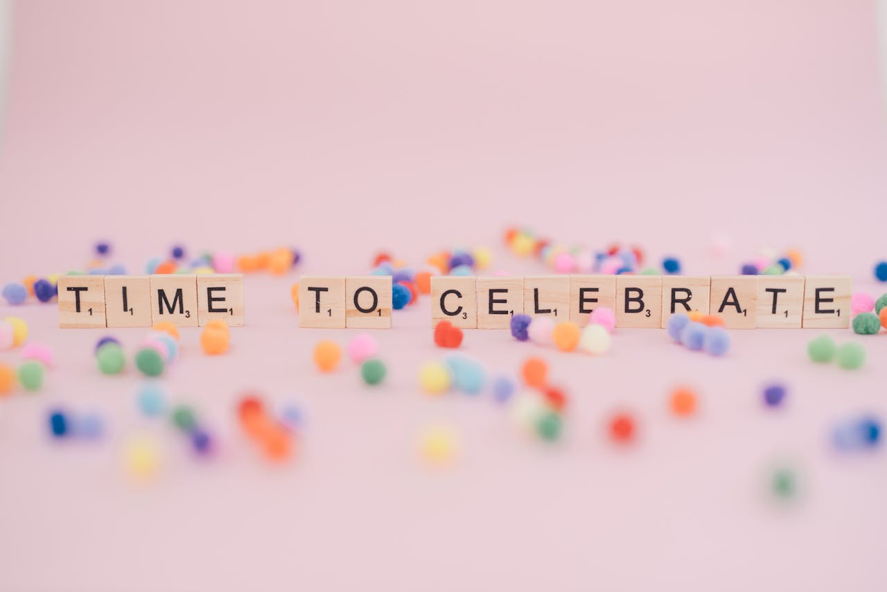 Colorful pom poms and alphabet tiles on pink background spelling 'Time to Celebrate.'